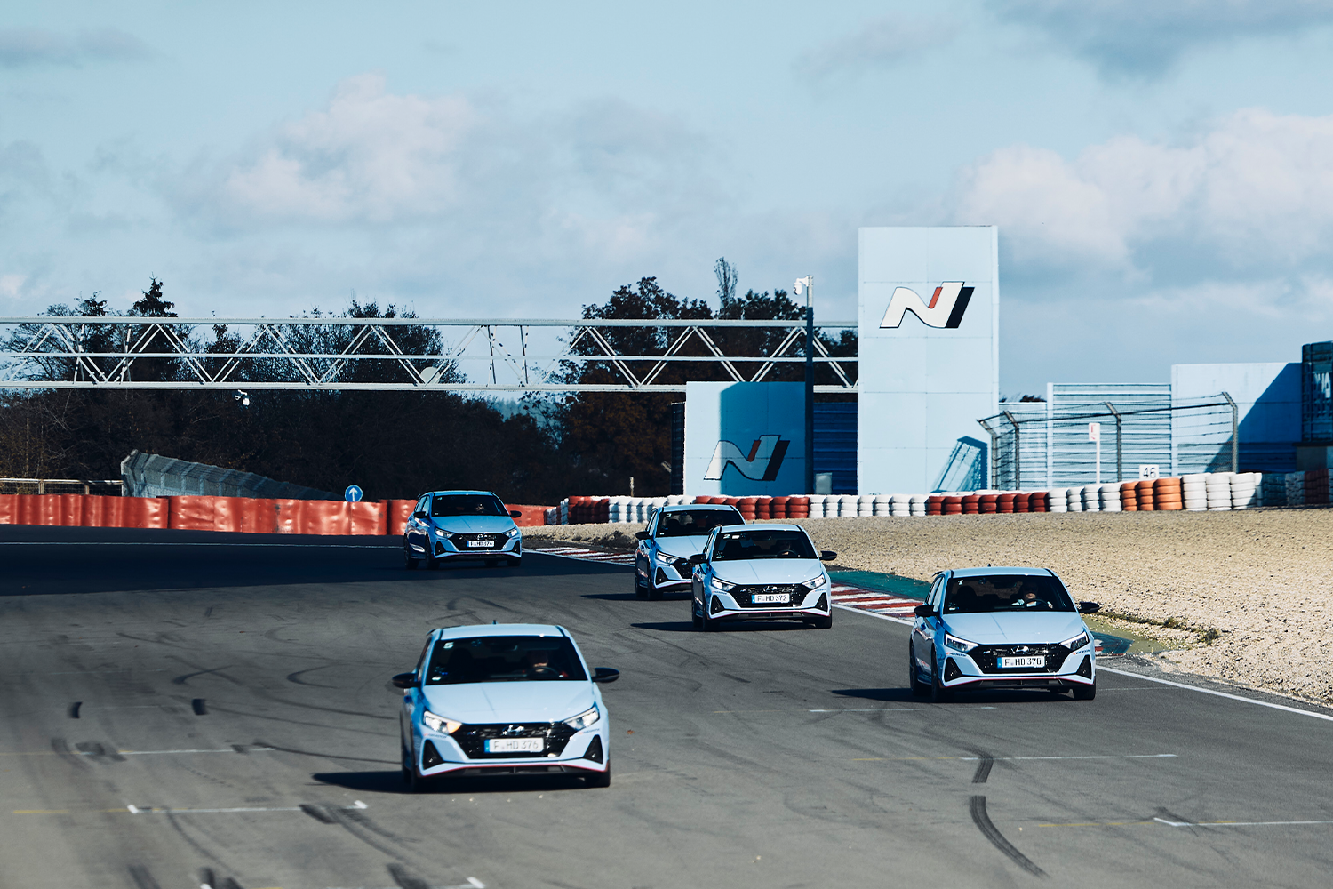A lineup of Hyundai i20 N cars driving on a race track during a training session. The cars are positioned in a staggered formation, showcasing dynamic handling. The background features the Hyundai N logo and safety barriers, emphasizing the motorsport setting under clear blue skies.
