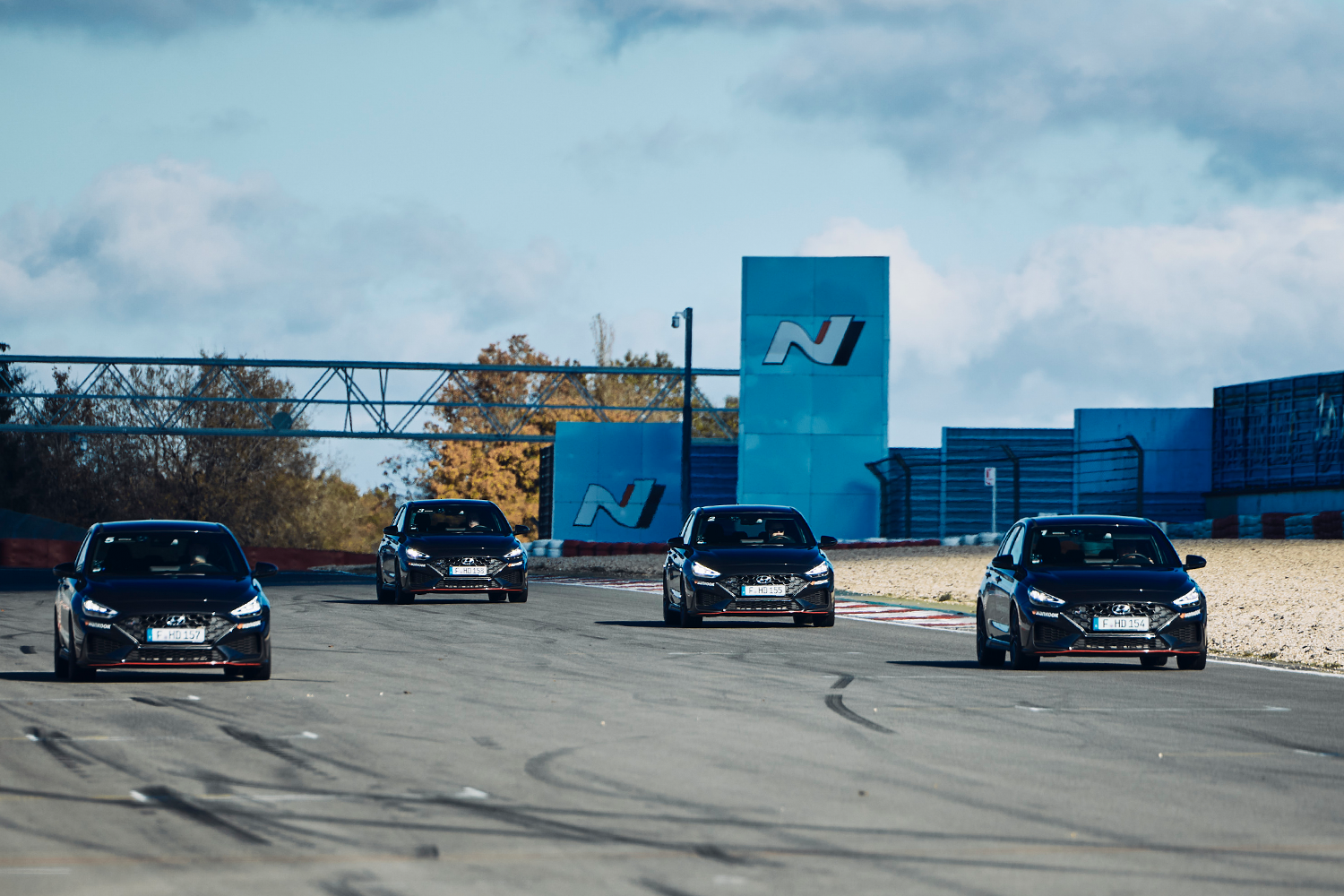 Four black Hyundai i30 N vehicles driving in a line on a race track under clear skies, with Hyundai's N branding visible in the background.
