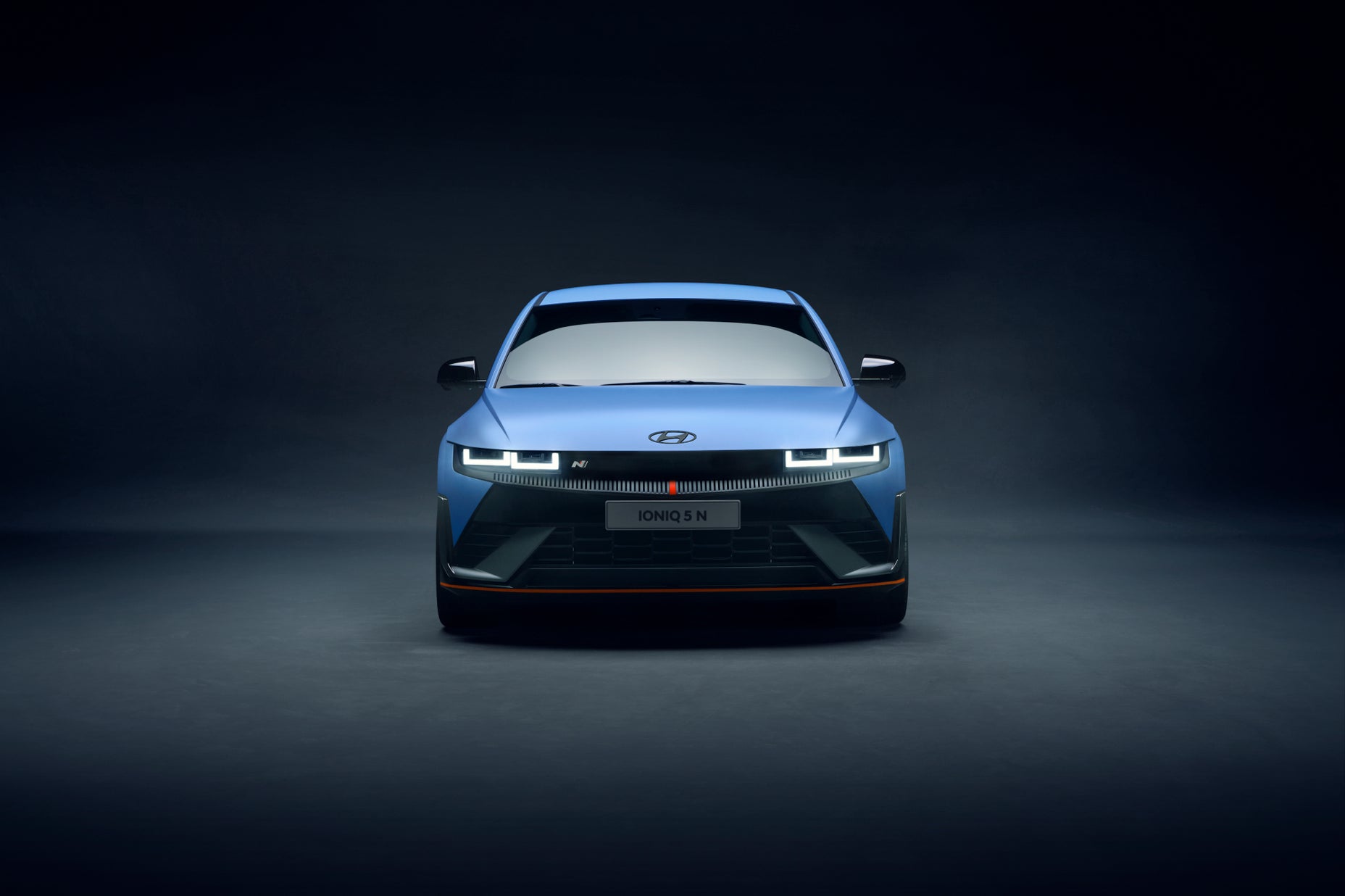 Front view of the Hyundai IONIQ 5 N showcased under studio lighting, emphasizing its bold grille, sharp LED headlights, and sporty stance against a dark background.
